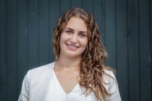 Mona Rakel. Frau mit dunkelblonden gelockten, schulterlangen Haaren lächelt freudestrahlend in die Kamera. Sie tägt eine helle Bluse mit Blazer vor dunklem Hintergrund.